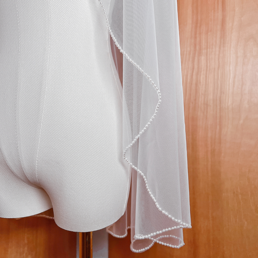 Close-up of a bridal veil featuring a dense cascading pearl trim along the edge, shown in ivory tulle at fingertip length.