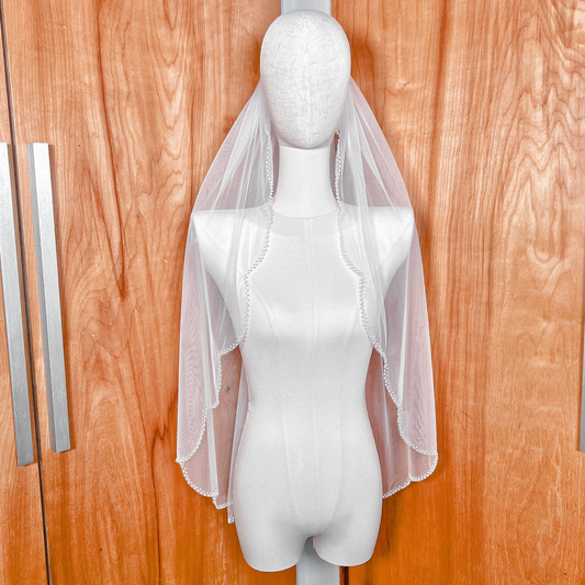 A single-tier ivory scalloped wedding veil with a delicate pearl edge shown in fingertip length.