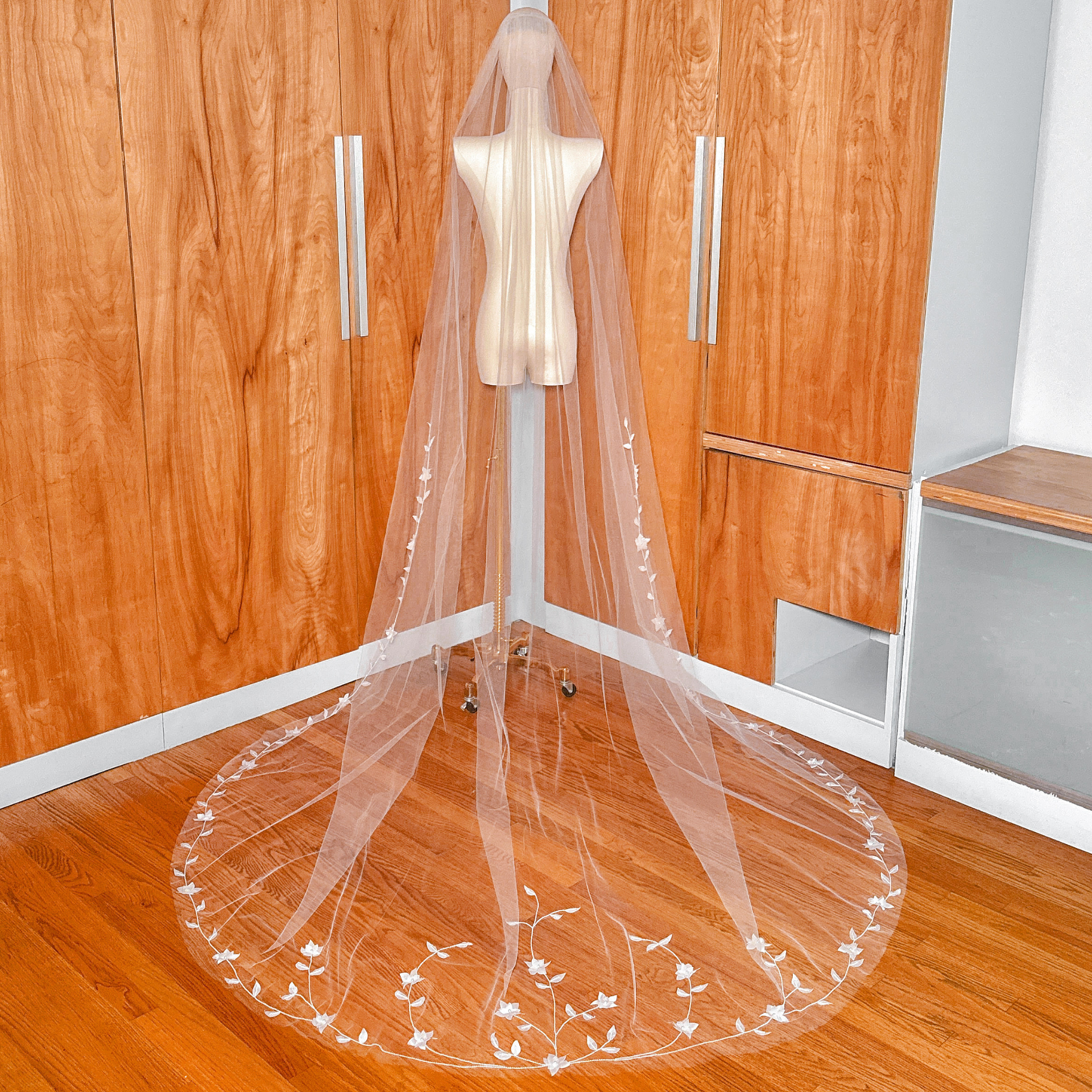 Full-length view of the sheer bridal veil draped gracefully on a mannequin, highlighting the overall silhouette and sweeping floral train against a wood backdrop.
