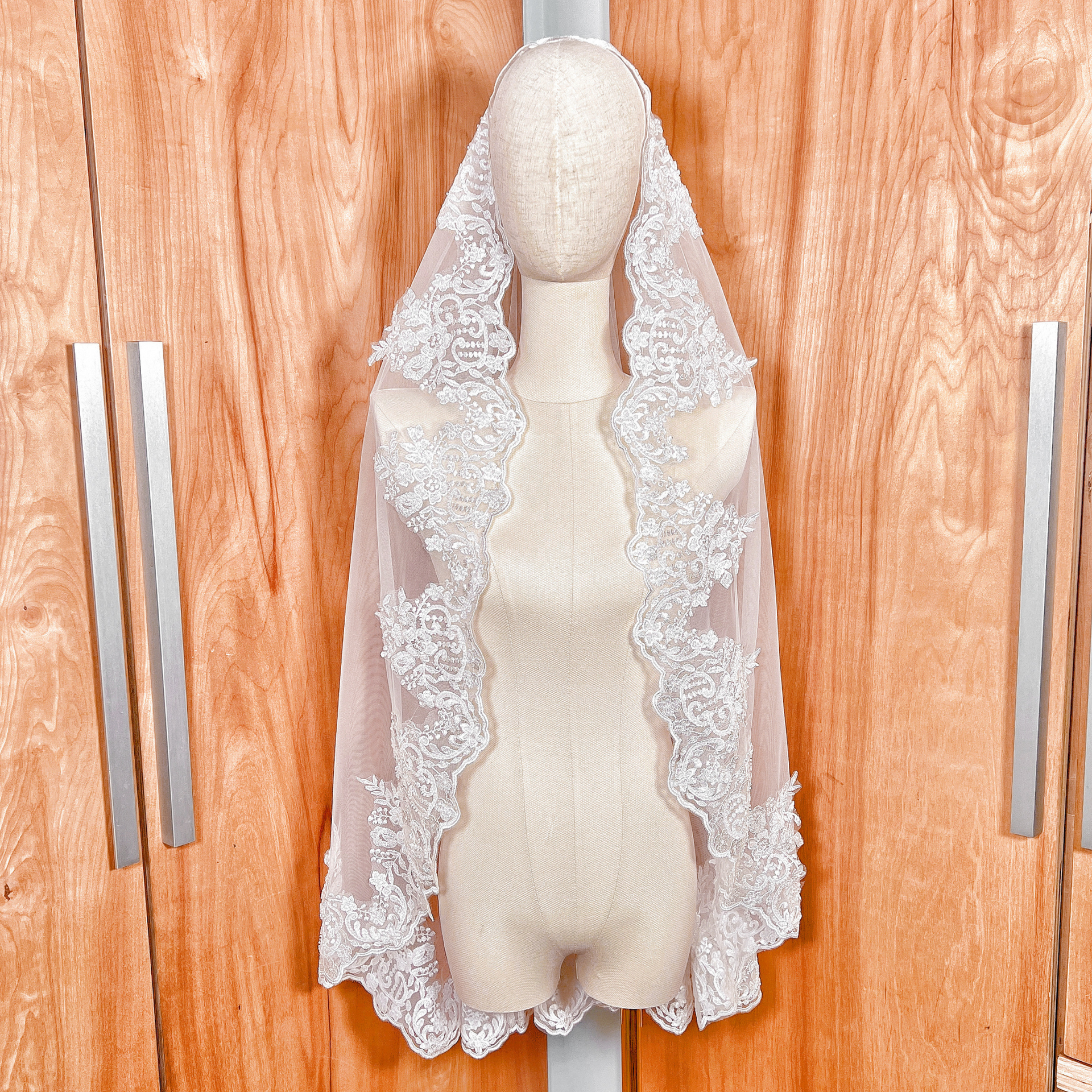 A fingertip length beaded lace mantilla wedding veil displayed on a mannequin, featuring intricate floral lace edging.