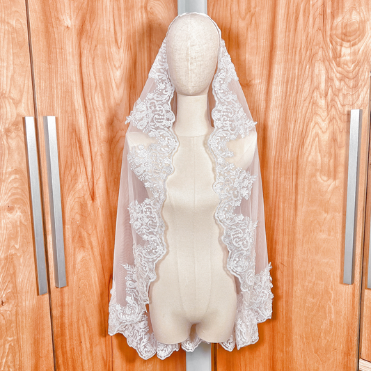 A fingertip length beaded lace mantilla wedding veil displayed on a mannequin, featuring intricate floral lace edging.
