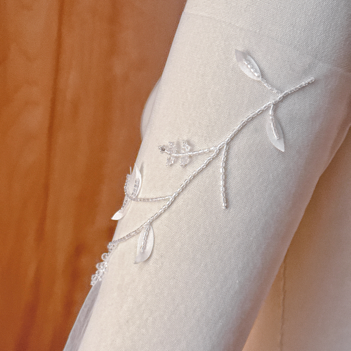 A close-up view of sheer illusion bridal sleeves featuring trailing vines hand-beaded with white seed beads and scattered 3D silk petals, displayed on a fabric dressmaker mannequin.