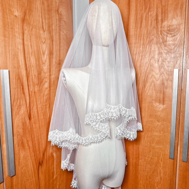 Elegant hip-length bridal drop veil with a delicate lace border, styled on a mannequin in a studio setting.