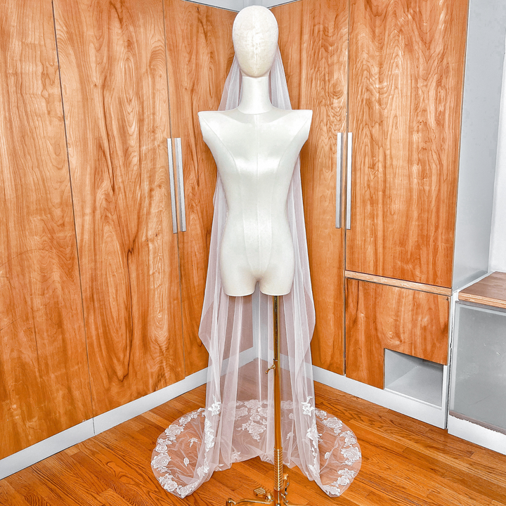 Soft white tulle of a floral lace edge wedding veil with long train cascading from a bridal mannequin.