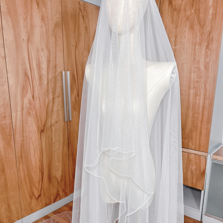 Elegant cathedral length wedding veil with two tiers and pearl trim, shown draped on a bridal form.