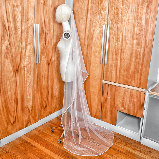 Side profile of a long mantilla wedding veil with ribbon edging draped over a mannequin's head and shoulders.