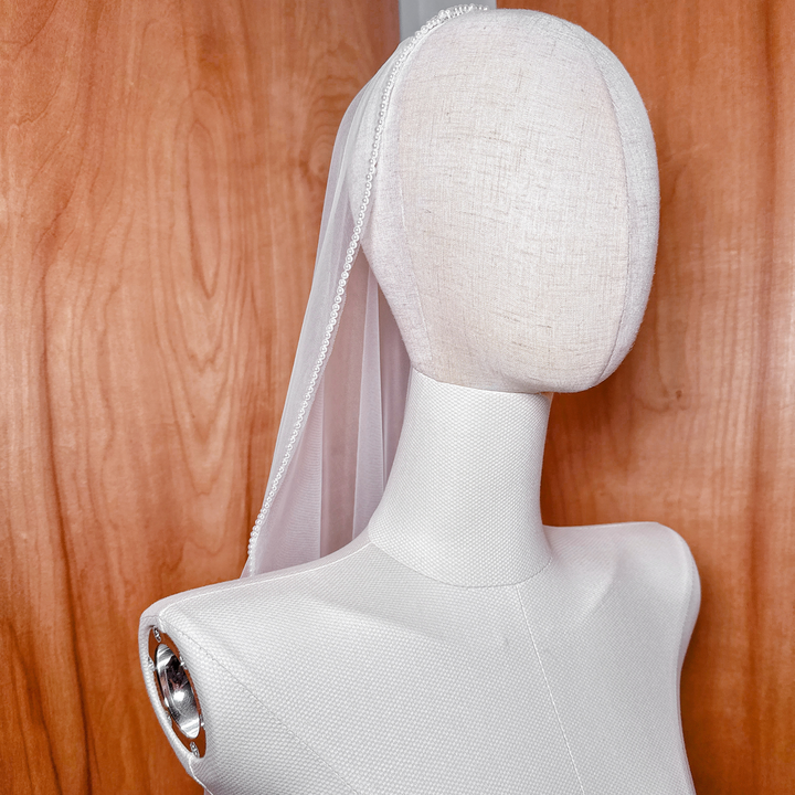 Detail shot of a pearl-embellished mantilla veil on a mannequin, showcasing the intricate pearl trim and soft ivory tulle.