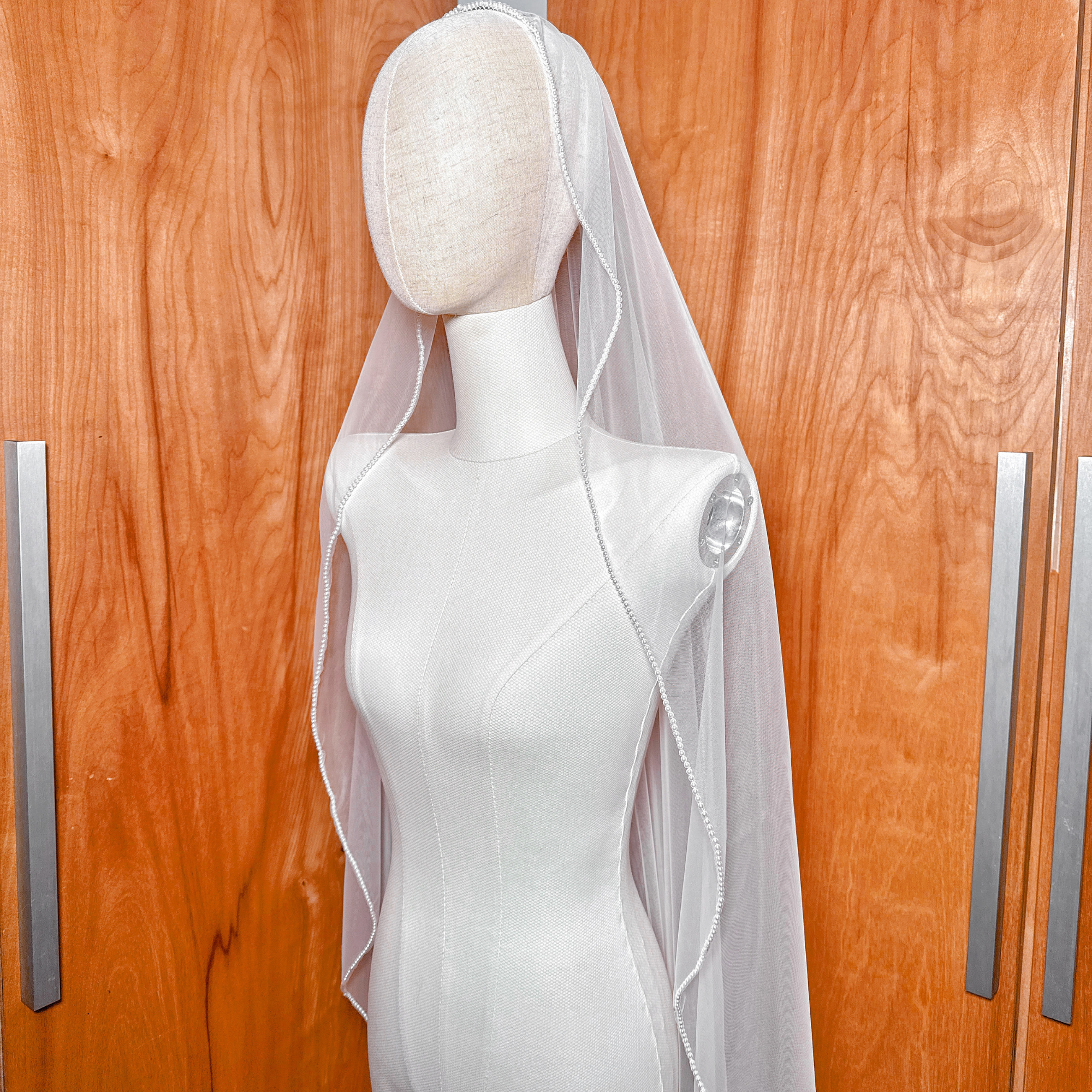 A waltz length wedding veil with a hand-stitched edge of pearls, shown on a mannequin to display the romantic, shimmering effect under studio lighting.