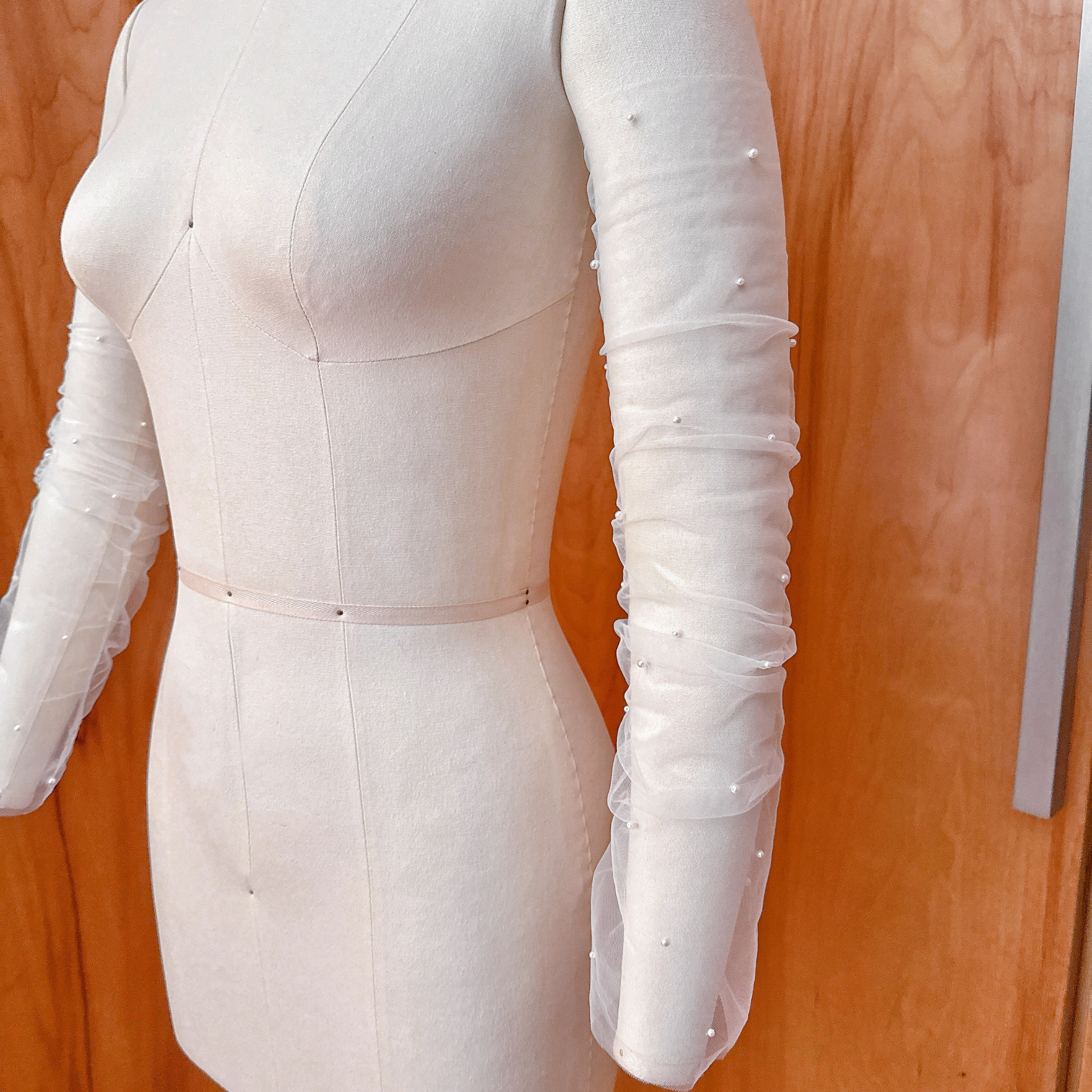 A close-up front view of sheer white ruched tulle detachable bridal sleeves embellished with scattered miniature pearls, displayed on a dressmaker mannequin.