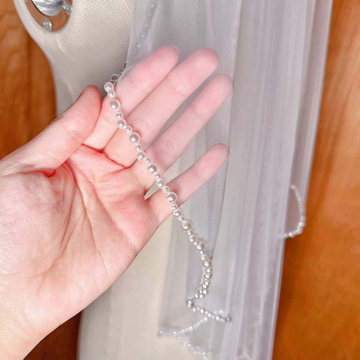 A close-up view of the delicate pearl trim along the edge of an elbow-length wedding veil, showing the intricate beadwork.