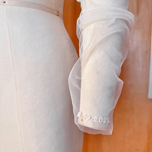 A close-up studio shot of a sheer ruched tulle bridal sleeve featuring a custom embroidered wedding date "8.19.2027 " stitched onto the delicate cuff area.